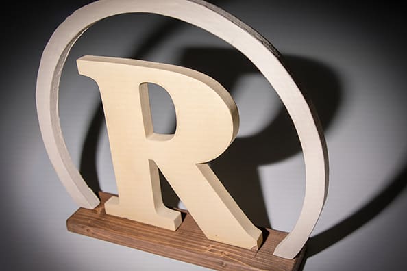 A wooden letter "R" is centered within a circular frame, showcasing its natural texture and warm colour