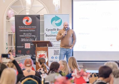 A Man stood on a stage holding a microphone talking to a room full of women sat around tables